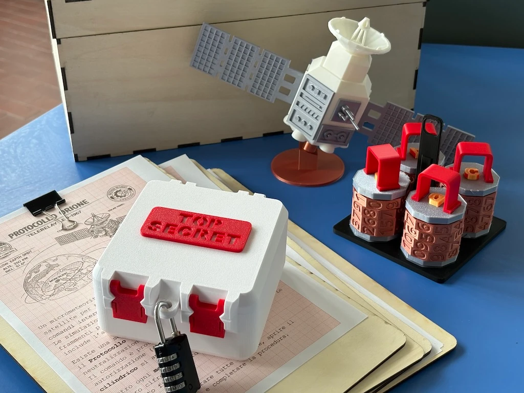 Prototipo fisico di escape didattico con scatola chiusa da lucchetto, modello di satellite, tre dispositivi e documenti di gioco.